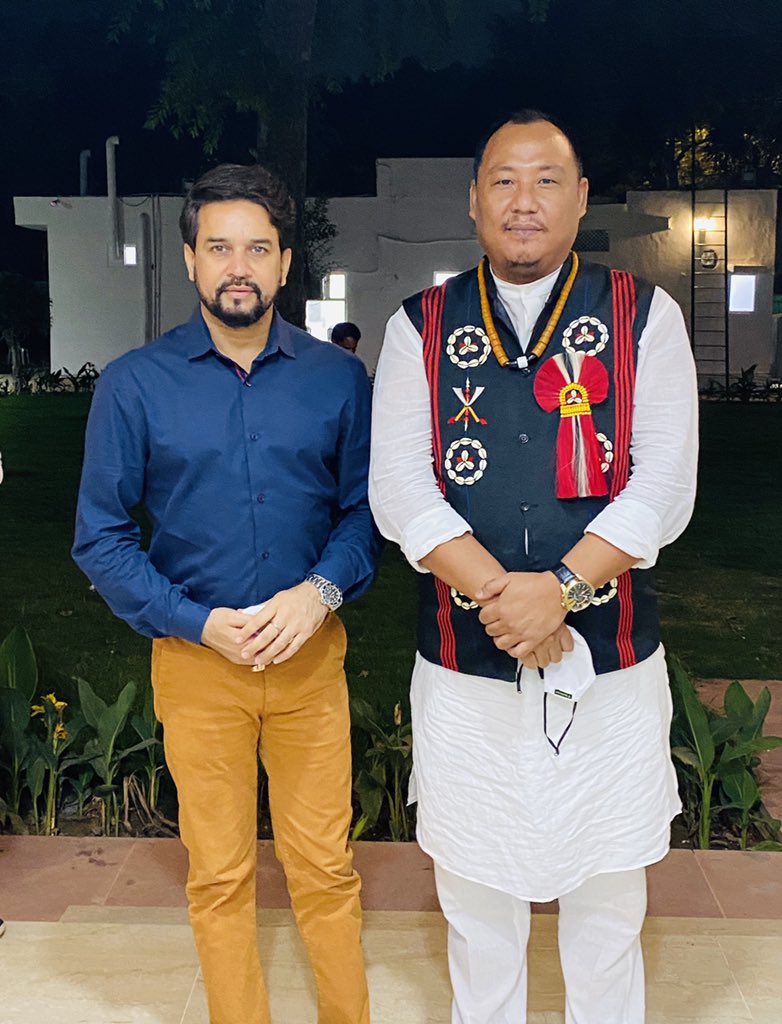 Benjamin Yepthomi standing in a traditional Naga waistcoat, bead necklace and white kurta at an evening engagement, photographed with another party leader.