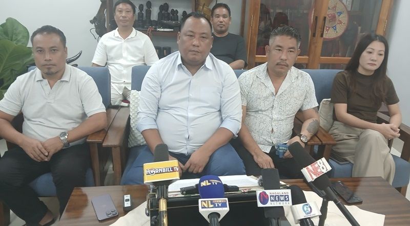 Benjamin Yepthomi seated at the centre of a press conference table with regional television microphones in front of him and party colleagues on either side.