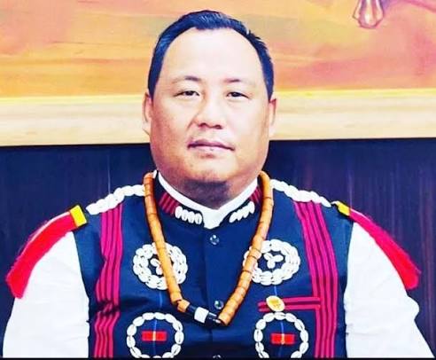 Benjamin Yepthomi in a traditional Naga waistcoat with cowrie-shell rosettes and an amber bead necklace, photographed indoors.