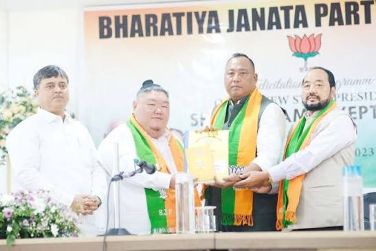 Benjamin Yepthomi receiving a memento at a felicitation programme on stage, flanked by three party colleagues wearing BJP scarves beneath a Bharatiya Janata Party banner.