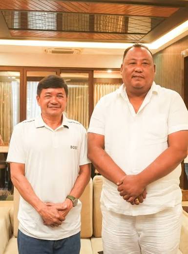 Benjamin Yepthomi standing beside a colleague in a wood-panelled interior, both dressed in white.