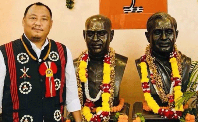 Benjamin Yepthomi in traditional Naga dress standing beside two garlanded memorial busts at a Bharatiya Janata Party office.